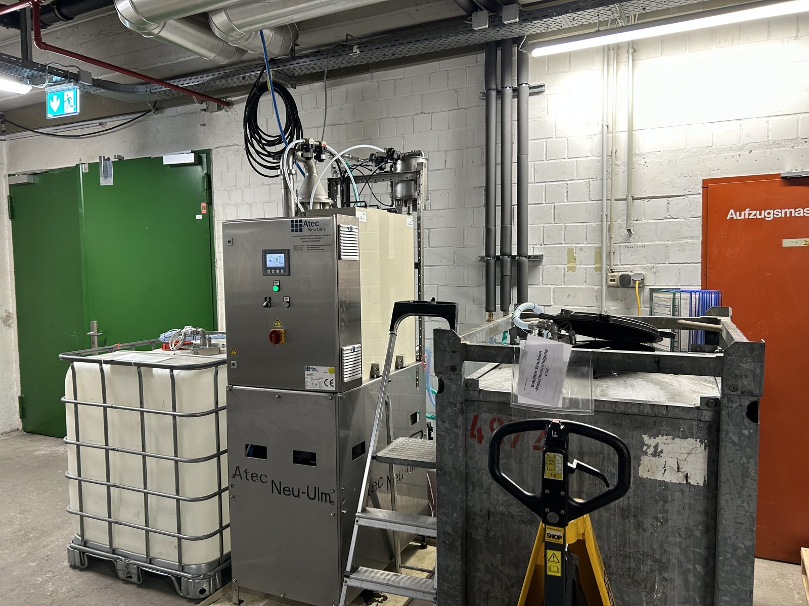 Atec Minitube in einem engen Technikraum mit gemauerten Wänden, daneben ein IBC-Behälter und ein Metallcontainer auf einem Hubwagen.