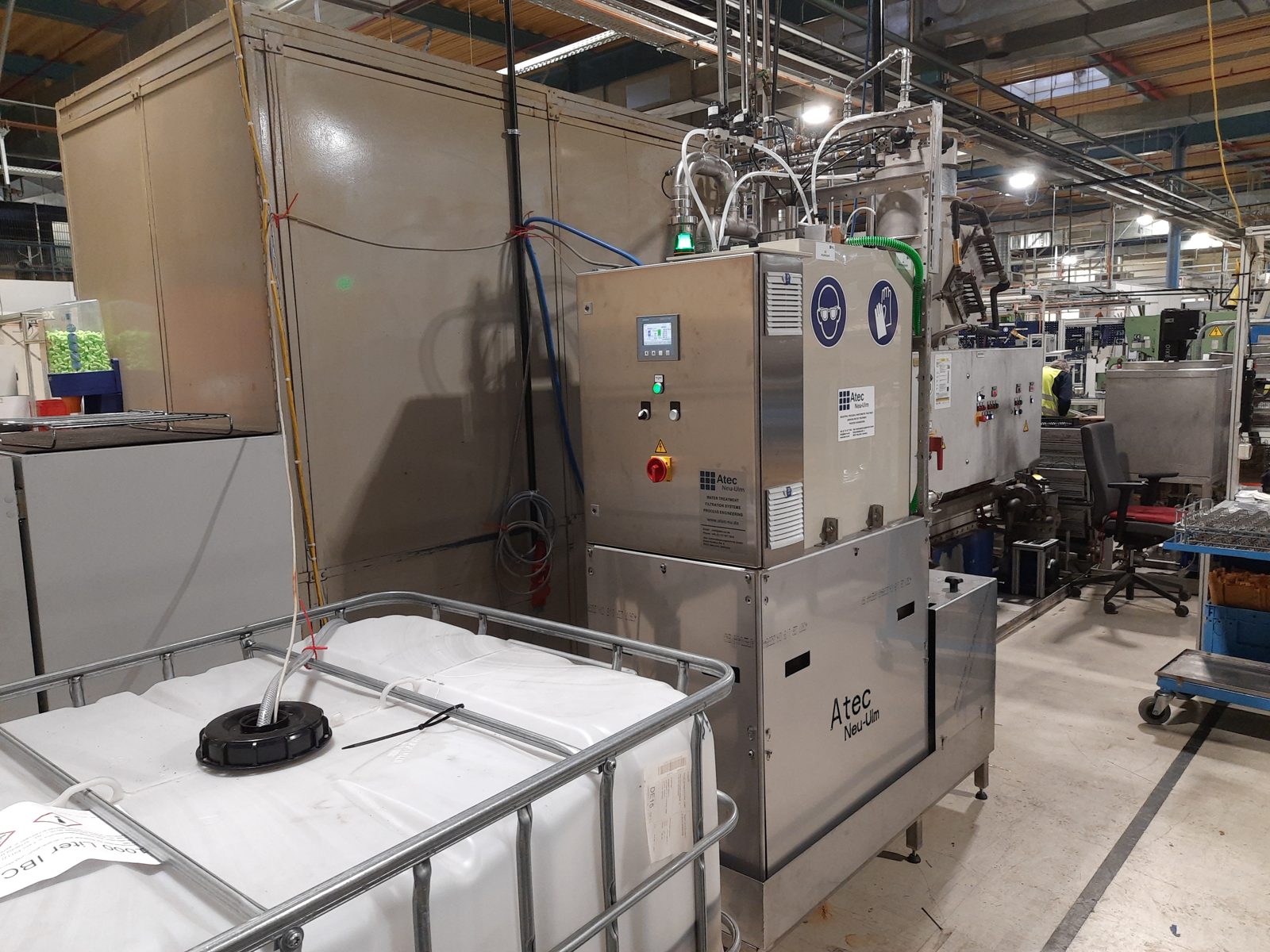 Atec Minitube aus Edelstahl mit Touch-Bedienpanel, aufgestellt direkt neben einer Fertigungsanlage in einer Produktionshalle; im Vordergrund ein IBC-Behälter.